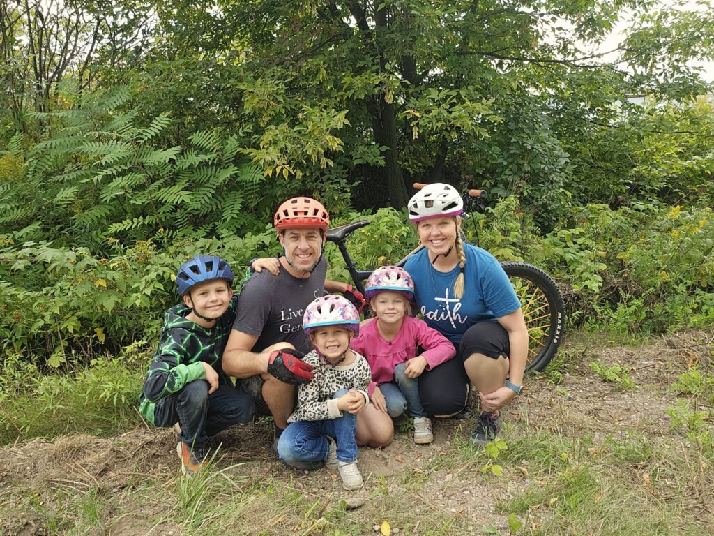 parents and three kids wearing bike helmets kneel posing in front of a bicycle in a wooded area
