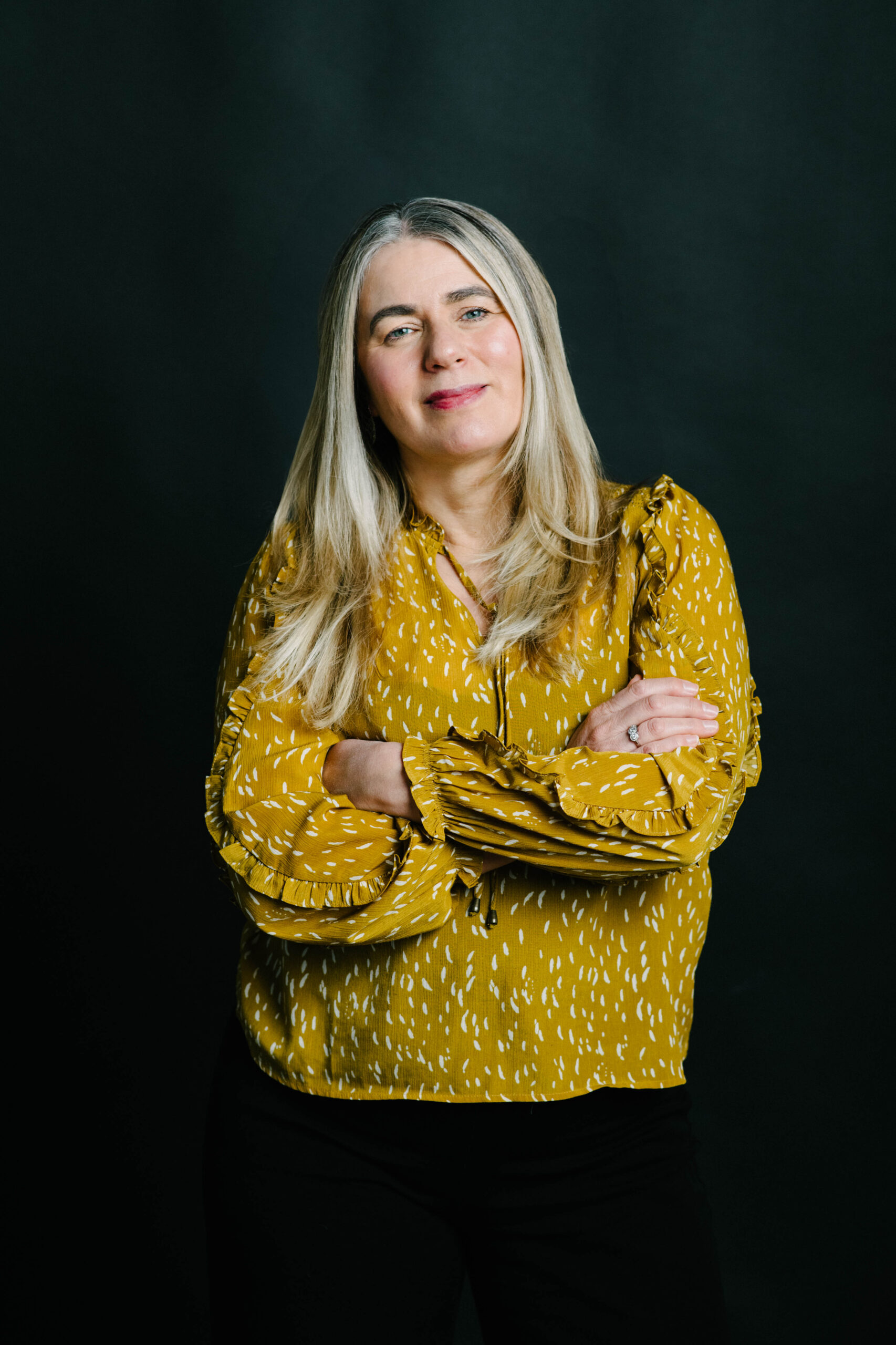 Dr. Guelay Bilen-Rosas standing with arms crossed, wearing a mustard yellow blouse against a dark background.